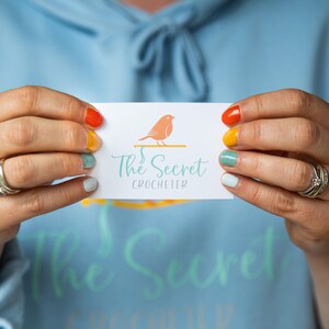 以下が含まれることがあります： 「The Secret Crocheter」の文字と、オレンジ色の鳥が枝にとまっているロゴが入った名刺。オレンジ、黄色、緑、青の色でペイントされた爪を持つ人がカードを持っています。背景は水色のパーカーです。