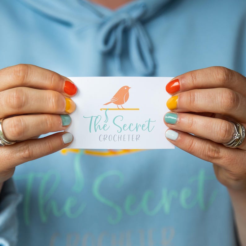 以下が含まれることがあります： 「The Secret Crocheter」の文字と、オレンジ色の鳥が枝にとまっているロゴが入った名刺。オレンジ、黄色、緑、青の色でペイントされた爪を持つ人がカードを持っています。背景は水色のパーカーです。
