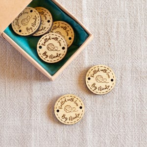 May include: Wooden craft tags with the text "Knitted with love By Nicola" and a yarn ball design. The tags are round with a stitched border and are in a box with a teal interior.