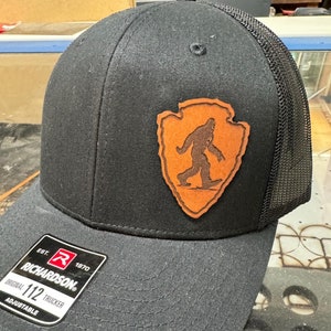 May include: Black trucker hat with a brown leather patch featuring a silhouette of a Bigfoot walking. The patch is shaped like a pointed arrowhead. The hat has a black mesh back and a black adjustable strap with the text "EST. 1970 RICHARDSON. ORIGINAL 112 TRUCKER ADJUSTABLE" printed on a white label.