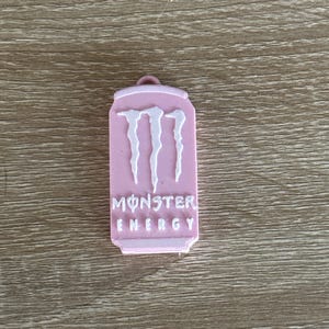 May include: A pink Monster Energy drink can-shaped keychain. The keychain features the Monster Energy logo in white, with the words "MONSTER ENERGY" printed below the logo. The keychain has a small loop at the top.