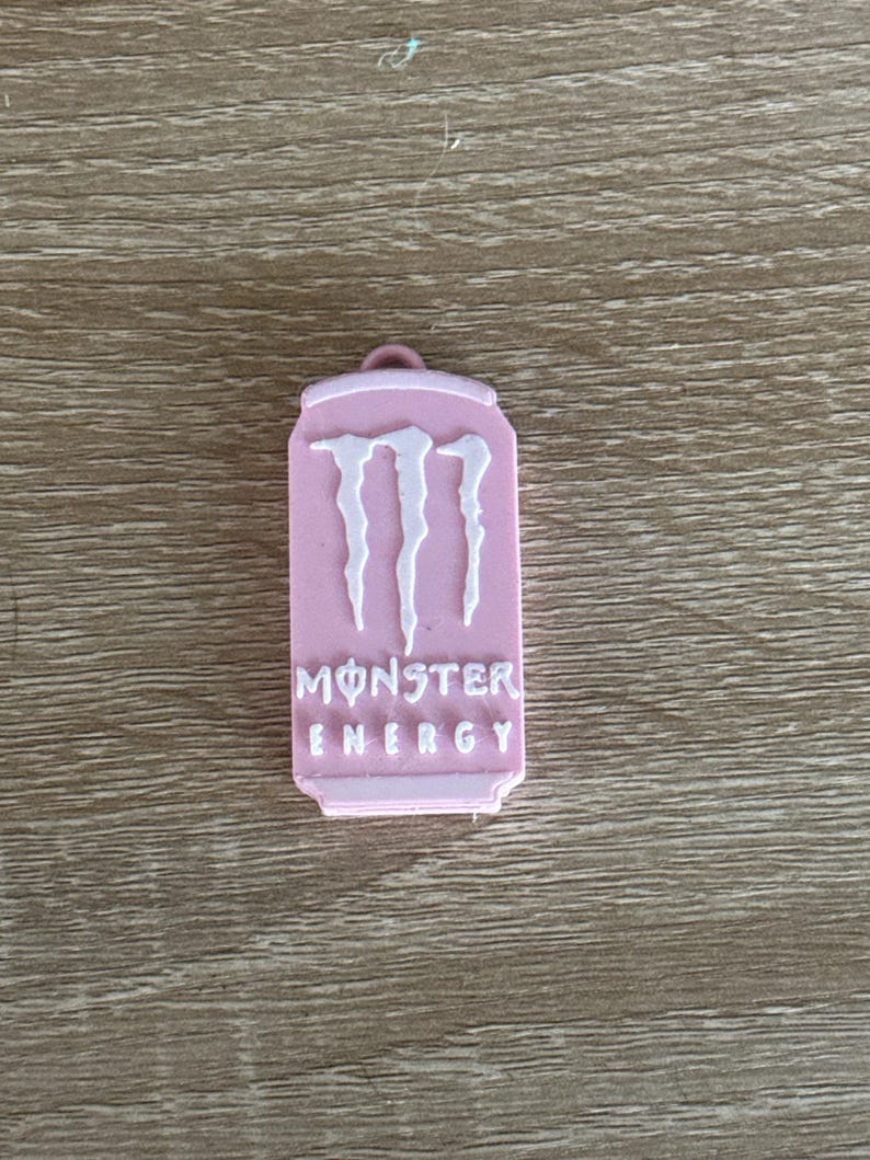 May include: A pink Monster Energy drink can-shaped keychain. The keychain features the Monster Energy logo in white, with the words "MONSTER ENERGY" printed below the logo. The keychain has a small loop at the top.