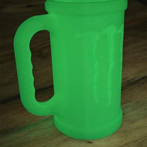 May include: A glowing green beer mug with a handle. The mug has a cylindrical body and a slightly flared rim. The handle is curved and attached to the side of the mug. The mug is sitting on a wooden surface.