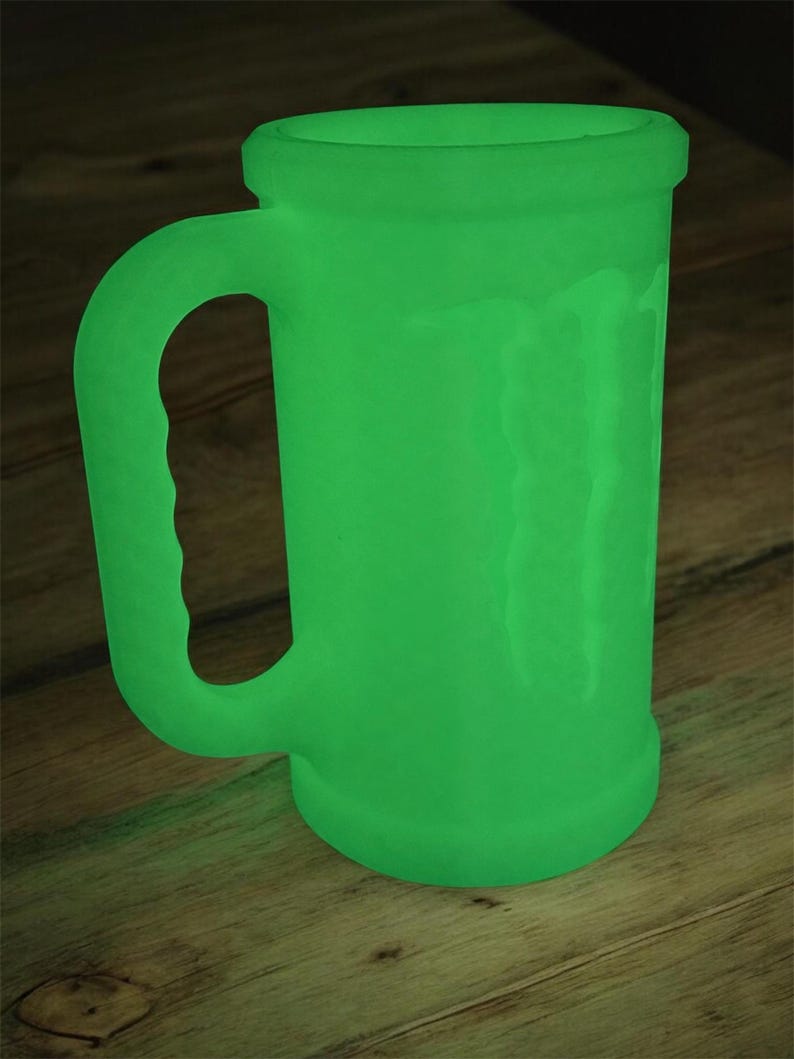 May include: A glowing green beer mug with a handle. The mug has a cylindrical body and a slightly flared rim. The handle is curved and attached to the side of the mug. The mug is sitting on a wooden surface.