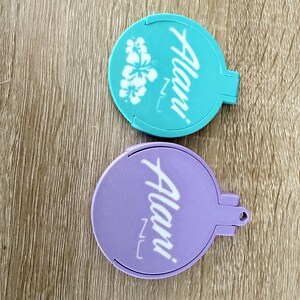 Puede incluir: Dos espejos compactos redondos, uno turquesa y otro lavanda. El espejo turquesa presenta un dise&ntilde;o floral blanco y la palabra "Alani" en escritura blanca. El espejo lavanda tiene la misma escritura. Ambos tienen un asa peque&ntilde;a.