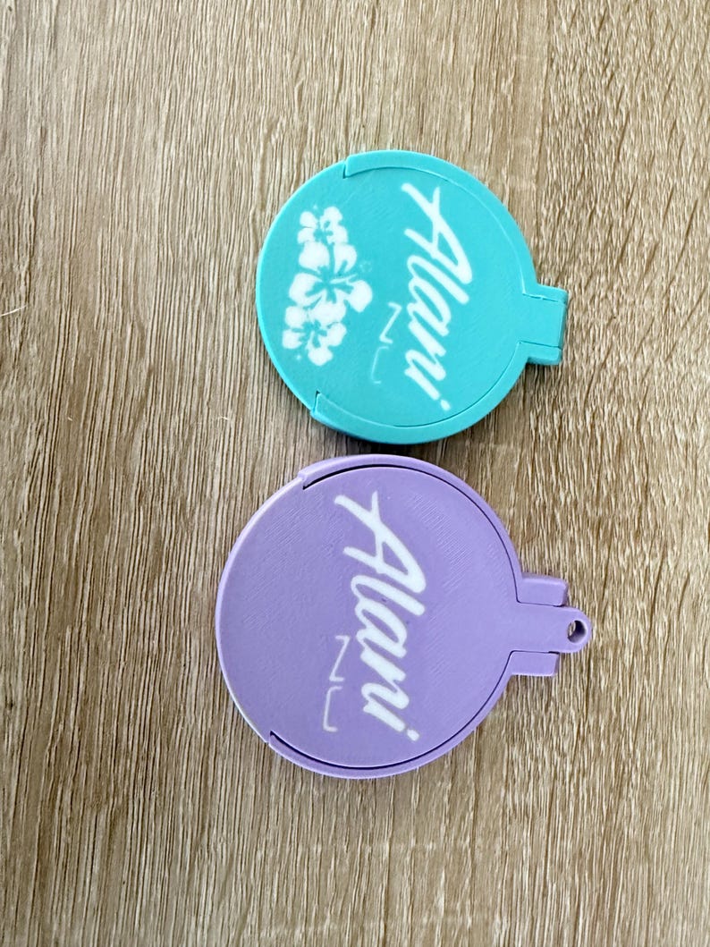 Puede incluir: Dos espejos compactos redondos, uno turquesa y otro lavanda. El espejo turquesa presenta un dise&ntilde;o floral blanco y la palabra "Alani" en escritura blanca. El espejo lavanda tiene la misma escritura. Ambos tienen un asa peque&ntilde;a.
