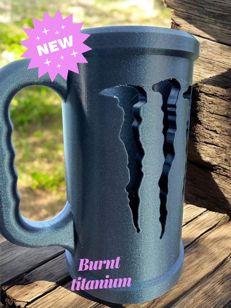 May include: A large, dark gray mug with a handle, featuring a cut-out design. The mug has the text "Burnt titanium" and a "NEW" sticker. The mug is sitting on a wooden surface.