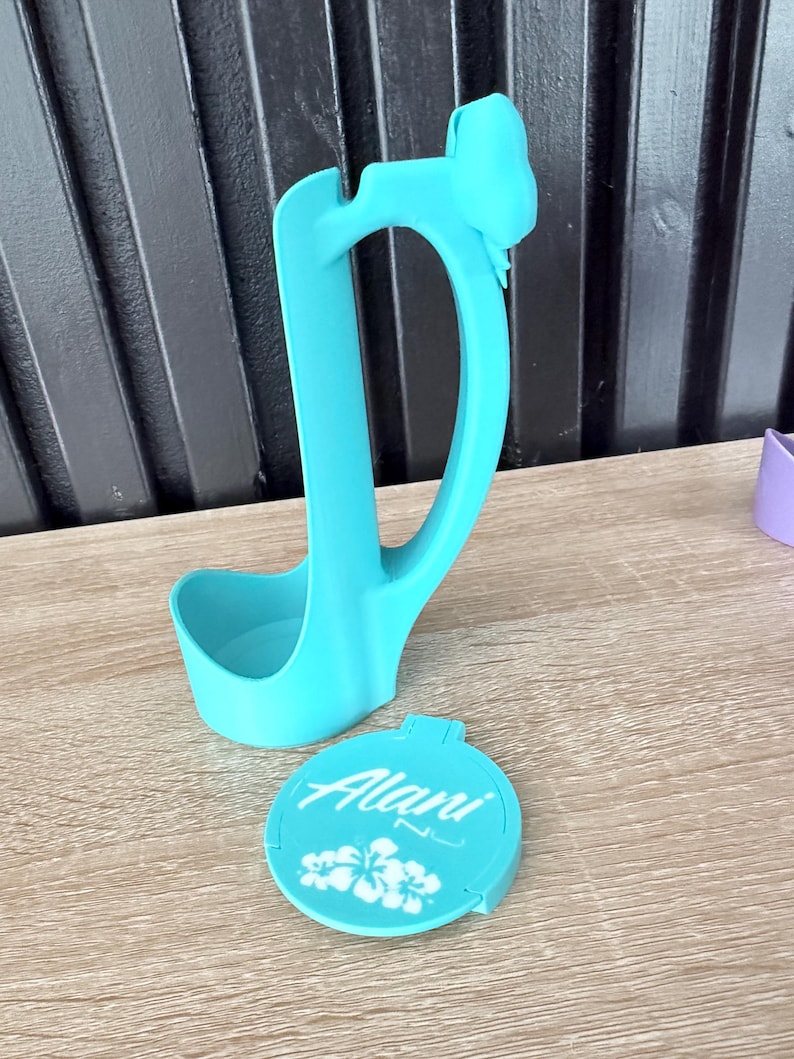 Puede incluir: Un cuchar&oacute;n azul claro con un asa en forma de lazo y una tapa a juego. La tapa es redonda y presenta el nombre "Alani" en escritura blanca, junto con dise&ntilde;os florales blancos. El cuchar&oacute;n y la tapa son de pl&aacute;stico.