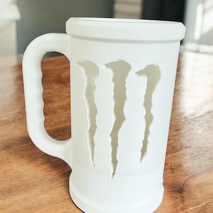 May include: A white ceramic beer stein with a handle. The stein features a cut-out design of three claw marks. The mug is cylindrical with a wide base and a thick rim. The mug is sitting on a wooden surface.