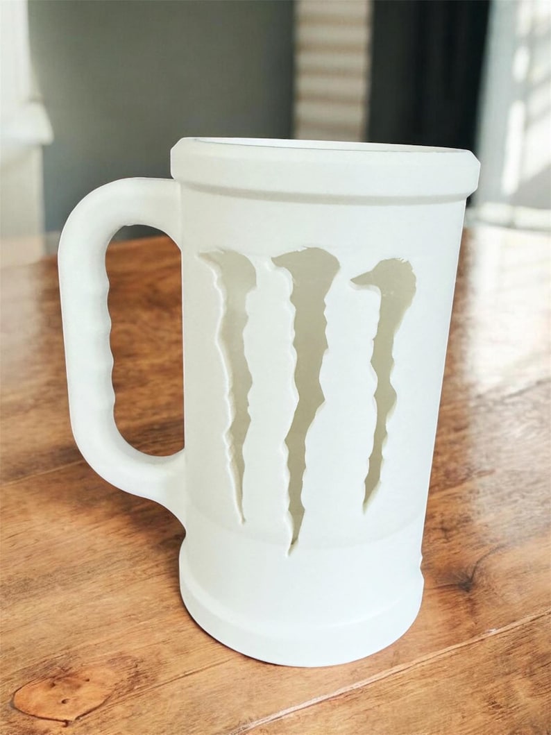 May include: A white ceramic beer stein with a handle. The stein features a cut-out design of three claw marks. The mug is cylindrical with a wide base and a thick rim. The mug is sitting on a wooden surface.