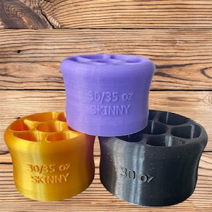 May include: Three colorful, cylindrical drink holders. One is gold, one is purple, and one is black. Each has a series of circular openings at the top and text that reads "30/35 oz SKINNY" or "30 oz".