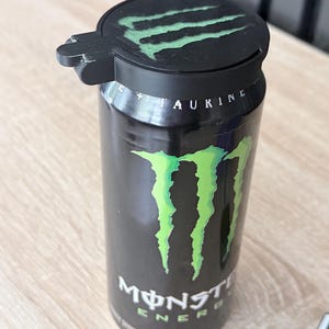 May include: A black Monster Energy drink can with a green claw logo and the word "Monster" in white. A black lid with a green claw logo is on top of the can. The can is on a light brown surface.