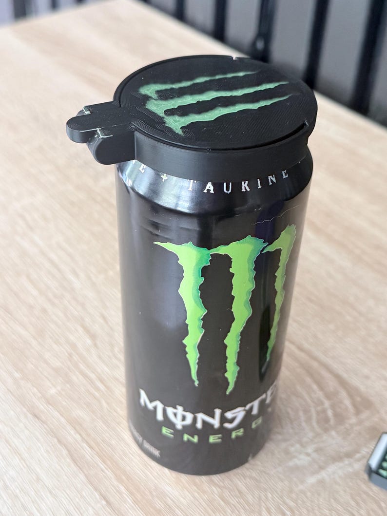 May include: A black Monster Energy drink can with a green claw logo and the word "Monster" in white. A black lid with a green claw logo is on top of the can. The can is on a light brown surface.