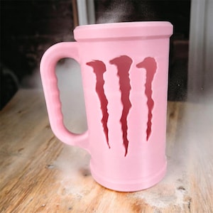 May include: A pink mug with a handle and three claw-shaped cutouts. The mug appears to be made of plastic and is sitting on a wooden surface. Steam rises from the top of the mug.