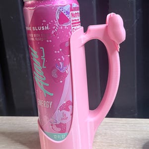 Puede incluir: Una lata rosa de 355 ml de bebida energ&eacute;tica "Pink Slush" en un soporte rosa a juego. La lata tiene un gr&aacute;fico de fresa y el texto "ENERGY" y "0g Sugar". El soporte tiene un asa y un lazo decorativo.