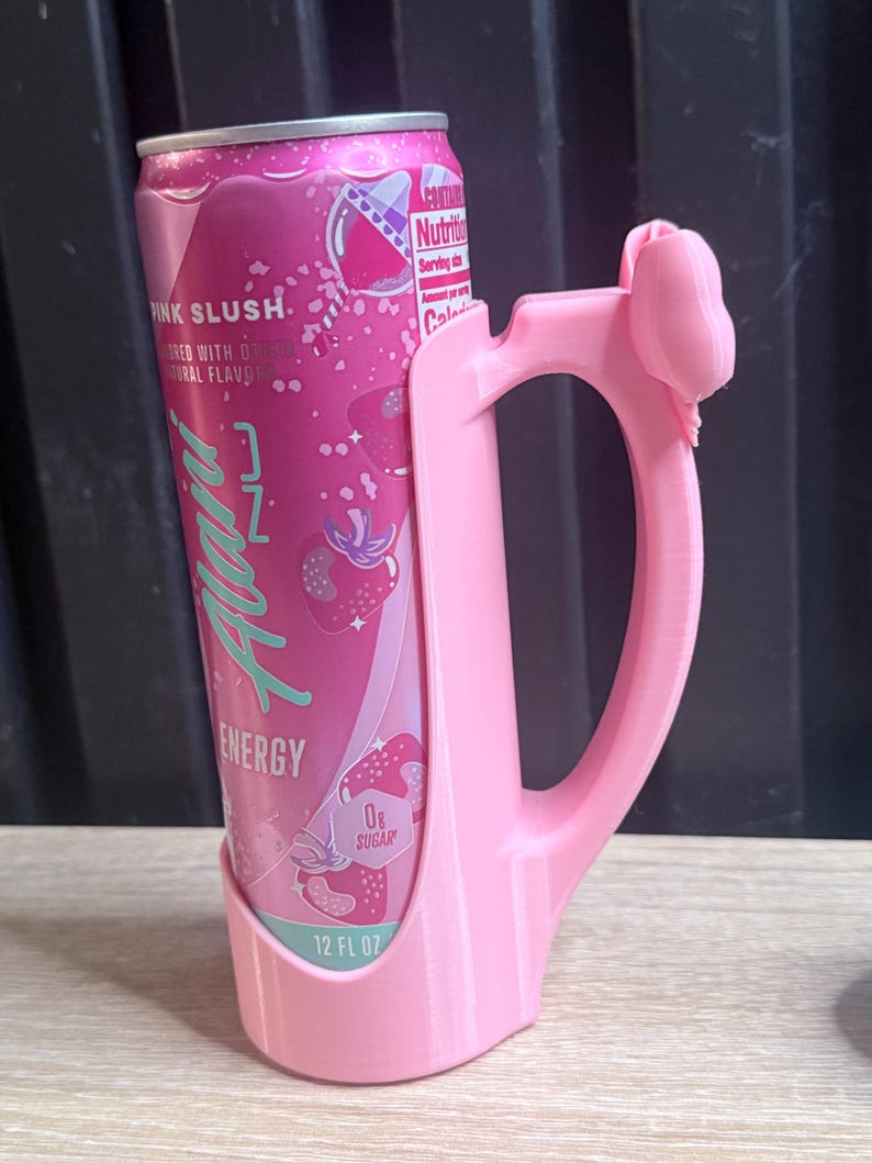 Puede incluir: Una lata rosa de 355 ml de bebida energ&eacute;tica "Pink Slush" en un soporte rosa a juego. La lata tiene un gr&aacute;fico de fresa y el texto "ENERGY" y "0g Sugar". El soporte tiene un asa y un lazo decorativo.