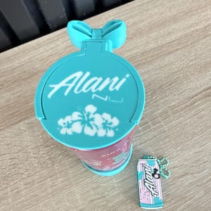 Puede incluir: Un vaso de color turquesa con la palabra "Alani" en escritura blanca, un asa en forma de lazo y un dise&ntilde;o floral. Tambi&eacute;n hay un llavero a juego con forma de lata.