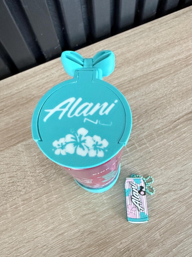 Puede incluir: Un vaso de color turquesa con la palabra "Alani" en escritura blanca, un asa en forma de lazo y un dise&ntilde;o floral. Tambi&eacute;n hay un llavero a juego con forma de lata.