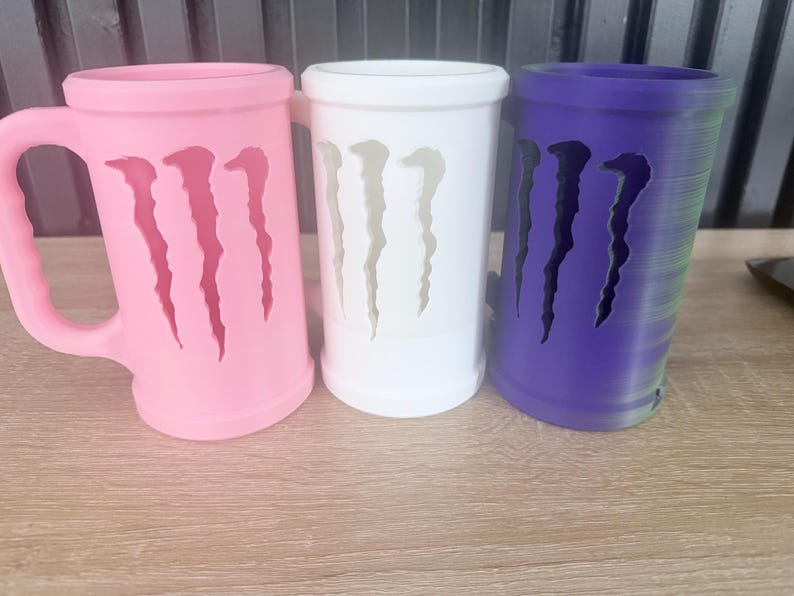 May include: Three colorful mugs with a distinctive design. The mugs are in pink, white, and purple. Each mug features a cut-out design resembling claw marks. The pink mug has a handle, while the others are cylindrical.