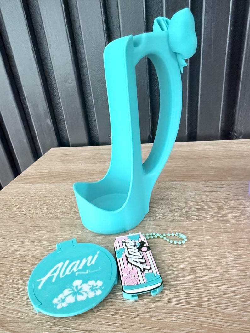 Puede incluir: Un portavasos de pl&aacute;stico azul claro con un detalle de lazo, un espejo compacto y un llavero. El espejo y el llavero tienen la palabra "Alani". El llavero tiene un dise&ntilde;o de rayas rosas y azules.