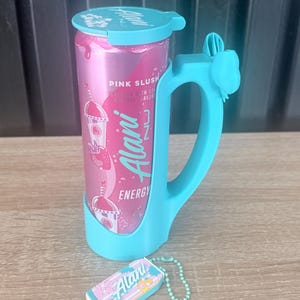 Puede incluir: Una lata rosa de bebida energ&eacute;tica Alani Nu con asa y tapa azules. La lata lleva la etiqueta "Pink Slush" y presenta un gr&aacute;fico de batido de fresa. Tambi&eacute;n hay un peque&ntilde;o llavero a juego con el logotipo de Alani Nu.