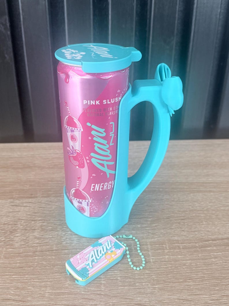 Puede incluir: Una lata rosa de bebida energ&eacute;tica Alani Nu con asa y tapa azules. La lata lleva la etiqueta "Pink Slush" y presenta un gr&aacute;fico de batido de fresa. Tambi&eacute;n hay un peque&ntilde;o llavero a juego con el logotipo de Alani Nu.
