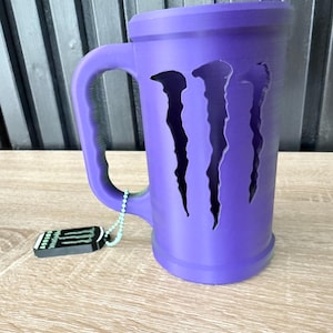 May include: A purple mug with a handle and the Monster Energy logo cut out on the front. A small tag with the Monster Energy logo hangs from the handle. The mug is sitting on a wooden surface.