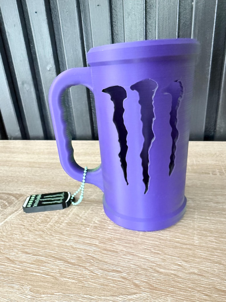 May include: A purple mug with a handle and the Monster Energy logo cut out on the front. A small tag with the Monster Energy logo hangs from the handle. The mug is sitting on a wooden surface.