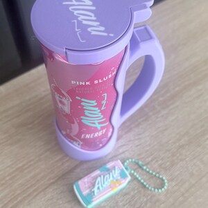Puede incluir: Un recipiente de bebida energ&eacute;tica Alani Nu rosa y morado con asa y llavero a juego. El recipiente tiene las palabras "Pink Slush" y "Energy" impresas. El llavero tiene el logotipo de Alani Nu.