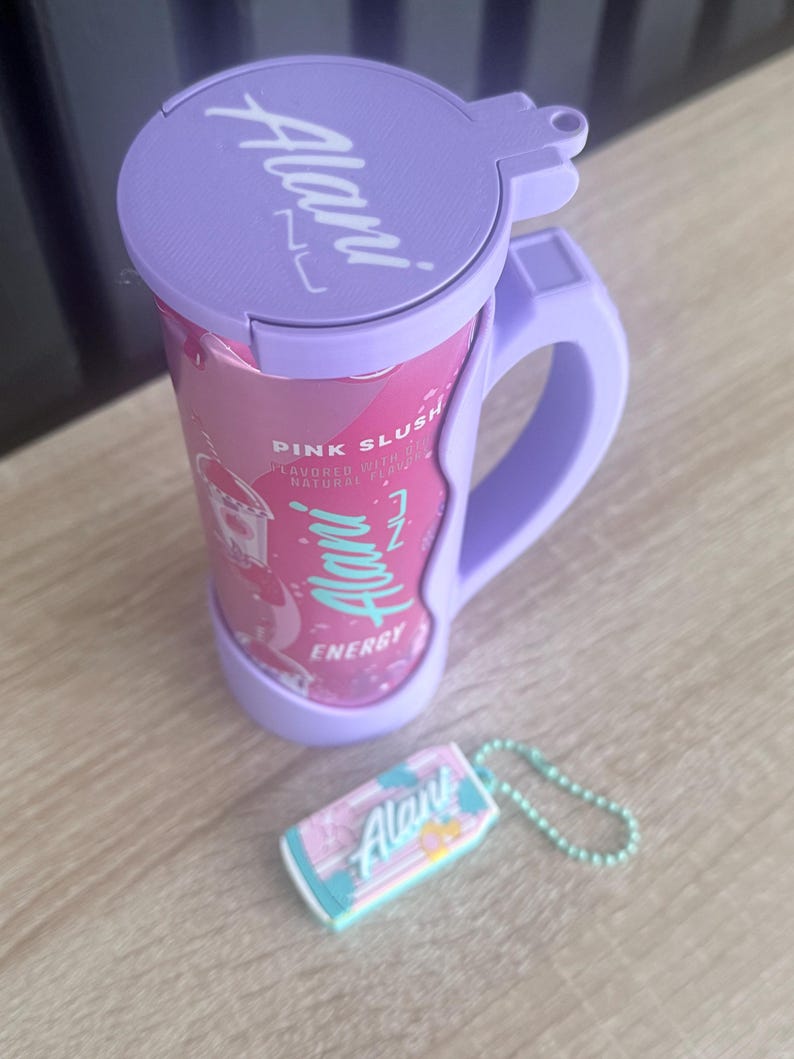 Puede incluir: Un recipiente de bebida energ&eacute;tica Alani Nu rosa y morado con asa y llavero a juego. El recipiente tiene las palabras "Pink Slush" y "Energy" impresas. El llavero tiene el logotipo de Alani Nu.