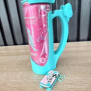 Puede incluir: Una lata rosa de bebida energ&eacute;tica con el texto "Pink Slush" y "Alani Energy" se sostiene en un soporte turquesa con asa y detalle de lazo. Un llavero a juego con el logotipo de Alani descansa cerca.