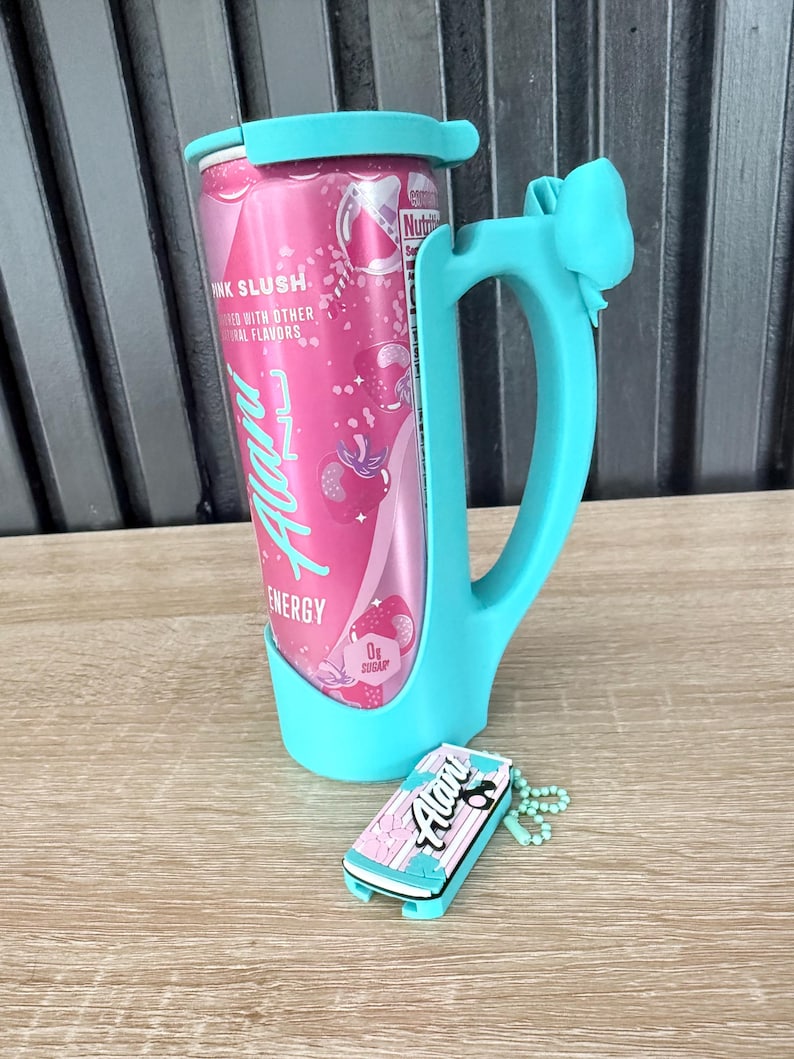 Puede incluir: Una lata rosa de bebida energ&eacute;tica con el texto "Pink Slush" y "Alani Energy" se sostiene en un soporte turquesa con asa y detalle de lazo. Un llavero a juego con el logotipo de Alani descansa cerca.