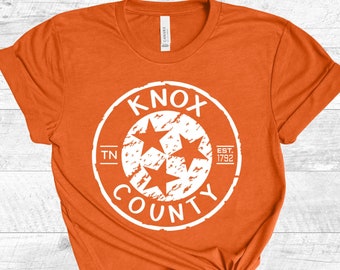 May include: Orange t-shirt with a white distressed circle graphic that says "Knox County" with three stars and "TN" and "Est. 1792" inside the circle.