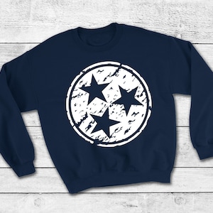 May include: A navy blue sweatshirt with a white distressed circle design featuring three stars. The stars are arranged in a triangular pattern.