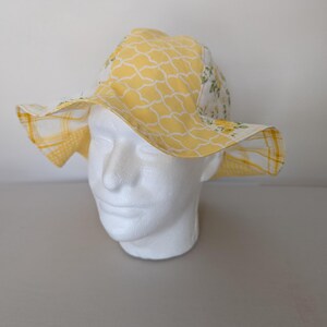 May include: A yellow and white patterned sun hat with a wide brim. The hat features a repeating geometric design and a floral pattern. The hat is displayed on a white mannequin head.