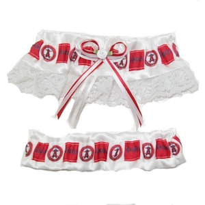 May include: A white satin garter set with red and white accents. The top garter features a lace trim and a red and white bow. Both garters are decorated with red rectangles with the word "ANGELS" and a logo.