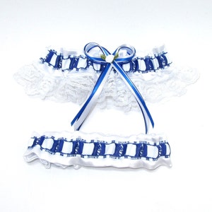 May include: A white satin garter set with blue and white checkered trim and a white lace edge. The top garter features a blue and white ribbon bow with a small white flower accent. The set is designed for a special occasion.