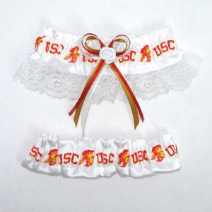 May include: White satin garter set with lace trim. The garters feature the letters "USC" in red and gold, along with a Trojan logo. A decorative bow with red and gold ribbons and a white rose embellishes the top garter.
