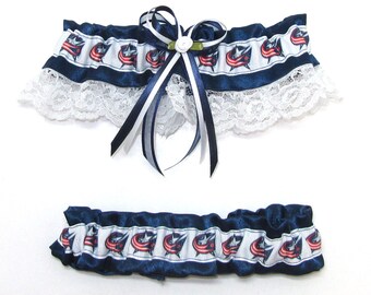 May include: Two-piece set of navy blue and white garters. The top garter features a white lace trim and a bow with navy and white ribbons. The garters have a repeating pattern of a red, white, and blue logo.