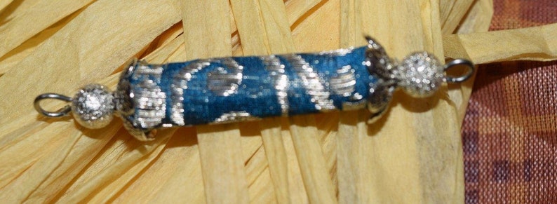May include: A small, silver-colored, decorative scroll with a blue fabric covering and silver beads at each end.