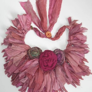 May include: A pink and red silk ribbon necklace with a large pink flower and two smaller flowers. The necklace is made of many layers of ribbon that have been frayed and tied together.