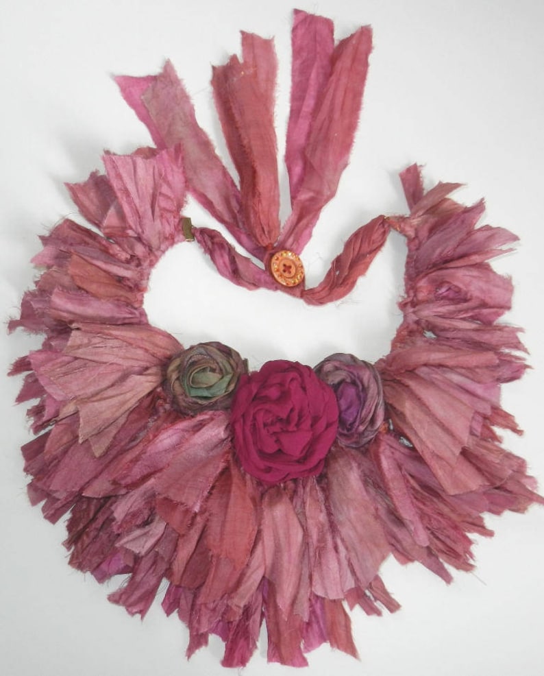 May include: A pink and red silk ribbon necklace with a large pink flower and two smaller flowers. The necklace is made of many layers of ribbon that have been frayed and tied together.