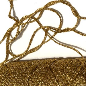 May include: A close-up of a small, rectangular gold-colored clutch with a matching gold cord strap. The clutch and strap are made of a woven, textured material, and the background is white.