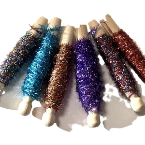 May include: A collection of colorful, glitter-covered wooden pegs. The pegs are adorned with shimmering tinsel in various colors, including blue, purple, brown, and red. The wooden ends are visible, creating a contrast with the vibrant tinsel.
