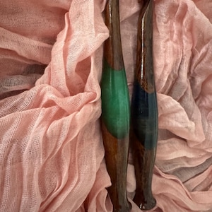 May include: Two handcrafted crochet hooks. The hooks have a dark brown wooden handle with a green and blue resin accent. The hooks are displayed on a pink, gauzy fabric background. The hooks are designed for yarn crafts.