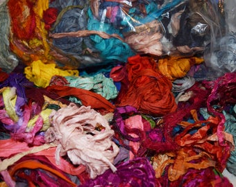 May include: A large plastic bag filled with colorful silk ribbon scraps. The ribbons are in various shades of red, orange, yellow, green, blue, purple, and pink. The ribbons are tangled and overlapping, creating a vibrant and textured image.