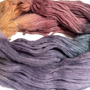 May include: A skein of hand-dyed yarn in a gradient of colors, transitioning from golden yellow to dusty rose, then to deep purple and finally to a muted blue-grey. The yarn appears soft and textured, ideal for knitting or crochet projects.