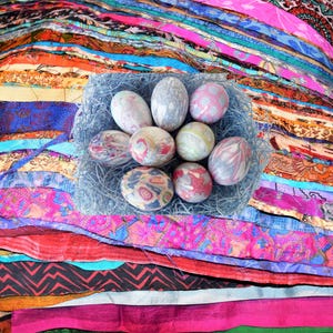 May include: A collection of decorative Easter eggs in a nest of blue-gray straw, set against a vibrant backdrop of layered, colorful fabric strips. The eggs feature various patterns and colors, creating a festive display.
