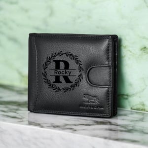 Puede incluir: Cartera de cuero negro con una inicial "R" monogramada dentro de una corona de laurel con el nombre "Rocky" debajo. La cartera tiene un cierre de solapa y un pequeño logotipo en la parte inferior.