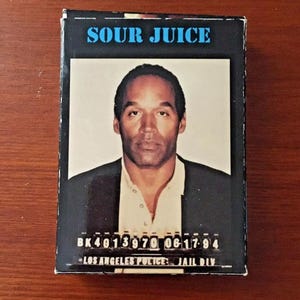 May include: A rectangular box with the words "SOUR JUICE" in blue letters at the top. The box features a portrait of a man in a suit and a white shirt. Below the portrait are the words "BK4013970 06-17-94 - LOS ANGELES POLICE: JAIL DIV."